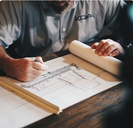 Dessinateur projeteur VRD au travail sur une table, en train de tracer au crayon le détail d'un plan technique de bâtiment, té gradué et rouleau de plans posés sur le bureau en bois